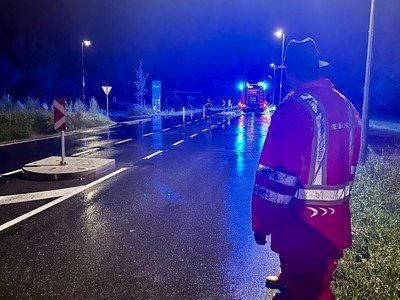Eine Person in einer reflektierenden Weste steht nachts auf einer nassen Straße und blickt auf Rettungsfahrzeuge mit blinkenden Lichtern. Straßenlaternen und ein Kreisverkehr sind im Hintergrund zu sehen.