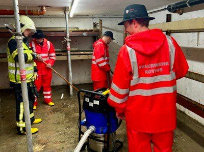 Vier Feuerwehrleute in roten Uniformen und Helmen arbeiten in einem Keller, einer hält einen Schlauch, ein anderer benutzt einen Staubsauger und zwei andere stehen daneben.
