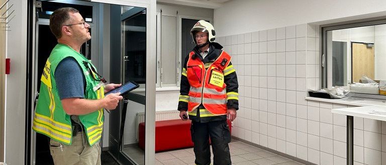 Ein Mann in einer grünen Sicherheitsweste hält ein Tablet und schaut einen Mann in einer Feuerwehruniform mit Helm und reflektierender Weste an. Sie stehen in einem Raum mit weißen Fliesen, einer roten Bank und einer Glastür.