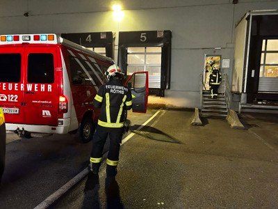 Ein Feuerwehrmann steht neben der offenen Tür eines roten Rettungswagens mit der Nummer 122. Ein anderer Feuerwehrmann betritt durch eine Tür ein Gebäude.