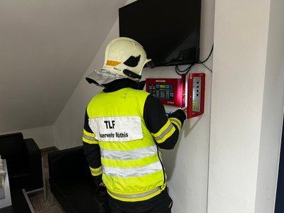 Ein Feuerwehrmann in einem gelben Vest installiert einen Feueralarm an einer weißen Wand. Der Feuerwehrmann trägt einen weißen Helm und eine reflektierende Weste. Es gibt eine schwarze Couch und ein Flachbildfernseher an der Wand befestigt.