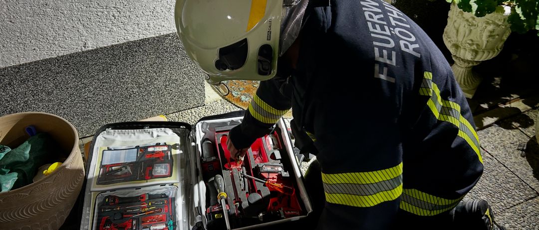 Ein Feuerwehrmann in voller Ausrüstung kniet auf dem Boden und arbeitet an einem Werkzeugkoffer mit verschiedenen Werkzeugen.
