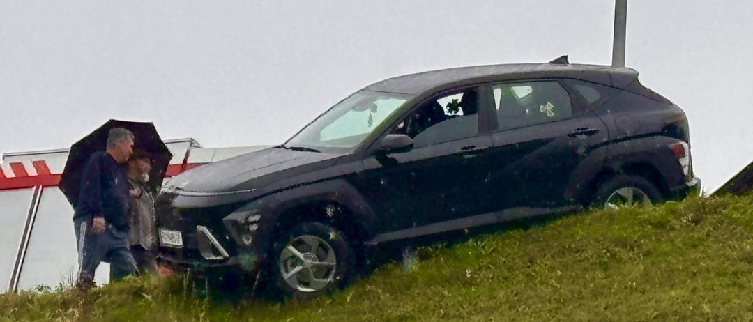 Ein schwarzer SUV ist auf einem Grashügel geneigt, mit einem Mann, der einen Regenschirm hält, und einer anderen Person in der Nähe.