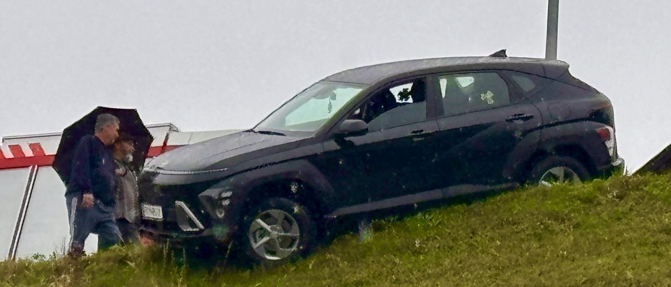 Ein schwarzer SUV ist auf einem Grashügel geneigt, mit einem Mann, der einen Regenschirm hält, und einer anderen Person in der Nähe.