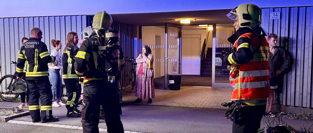 Feuerwehrleute in voller Ausrüstung stehen vor einem Gebäudeeingang, eine Frau und Zuschauer sind in der Nähe.