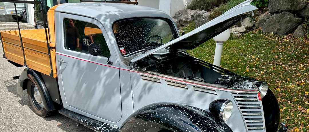 Ein alter grauer LKW mit geöffneter Motorhaube, am Straßenrand geparkt, mit einer Person im Inneren.