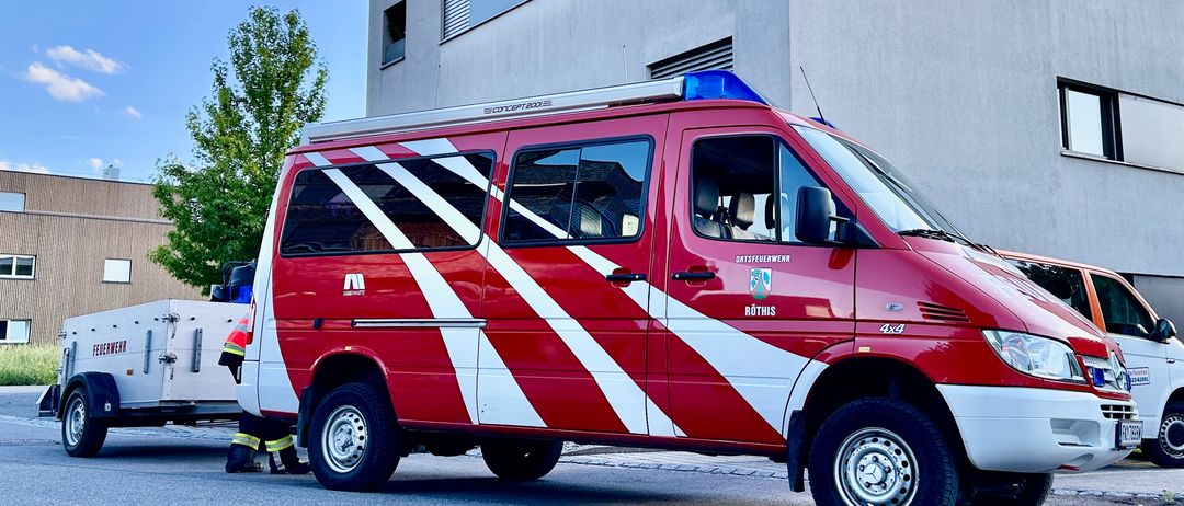 Ein rot-weißer Feuerwehrwagen mit dem Schriftzug 'Rotis' steht auf einer Straße mit einem Gebäude und einem Baum im Hintergrund.