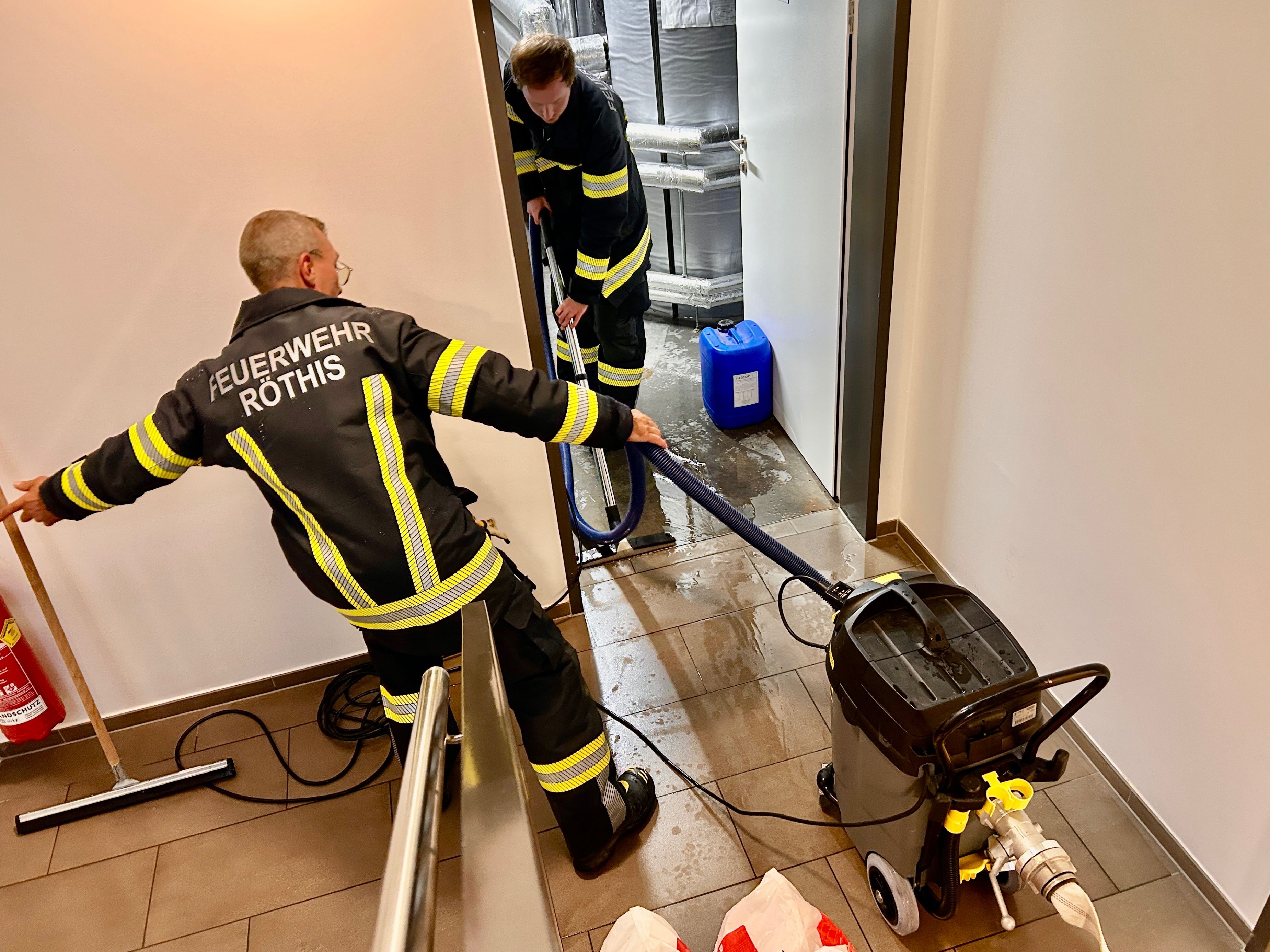 Zwei Feuerwehrleute benutzen einen Staubsauger, um Wasser aus einem überfluteten Korridor zu entfernen. Ein Feuerwehrmann hält den Staubsauger, während der andere den Schlauch hält.