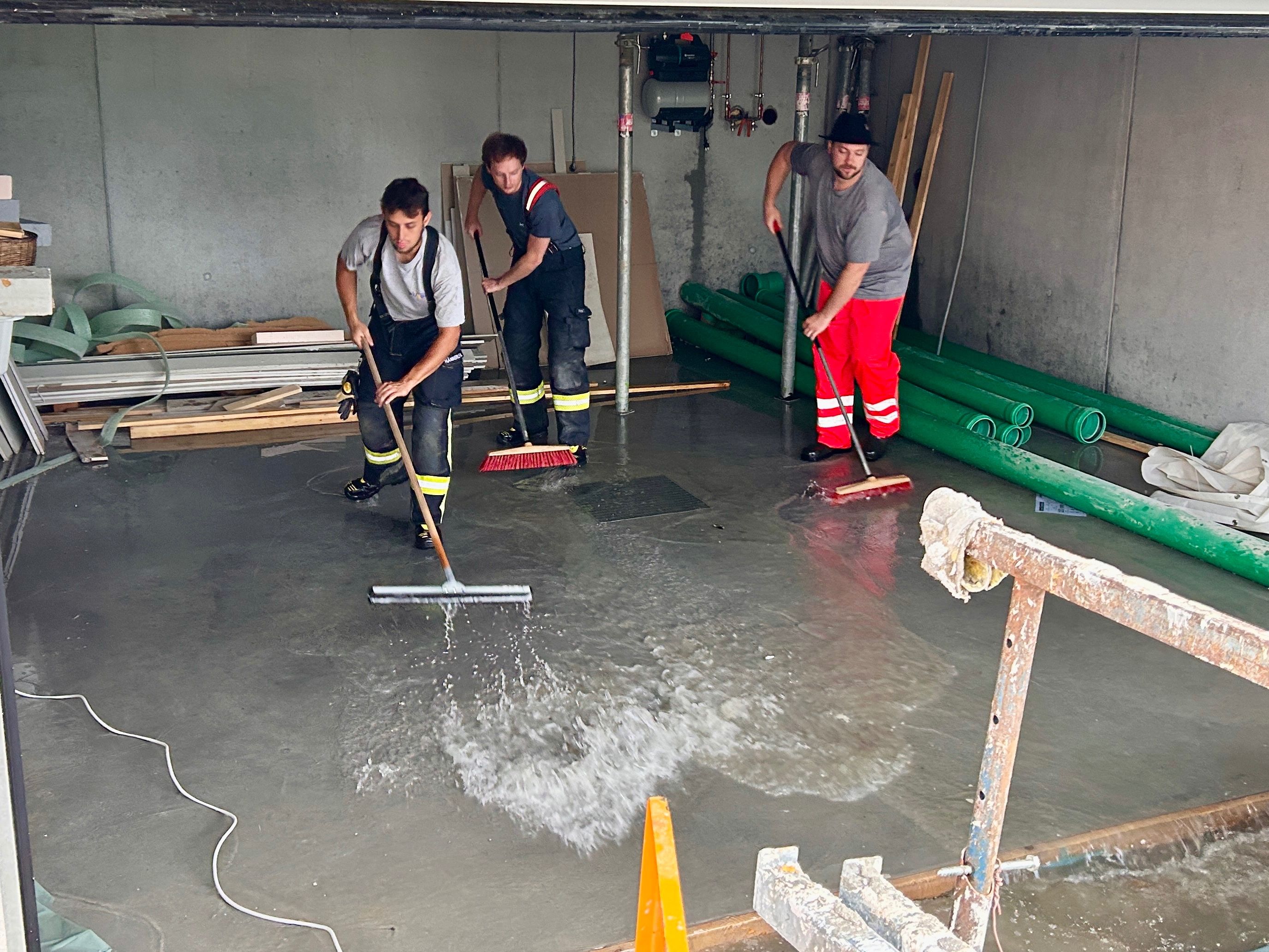 Drei Männer reinigen einen nassen Boden mit Besen. Einer hält einen Besen mit spritzendem Wasser. Ein weiterer trägt einen Hut und der dritte ist in reflektierender Kleidung. Der Hintergrund besteht aus einer Betonwand und grünen Rohren.