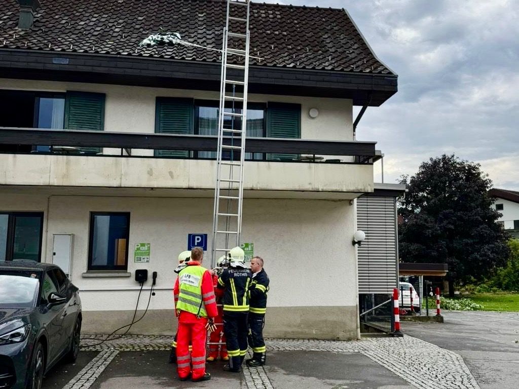 Feuerwehrleute und Rettungskräfte in Schutzkleidung stehen vor einem Gebäude, an dem eine Leiter zum Dach führt. Der Bereich ist mit roten und weißen Pollern abgesperrt.