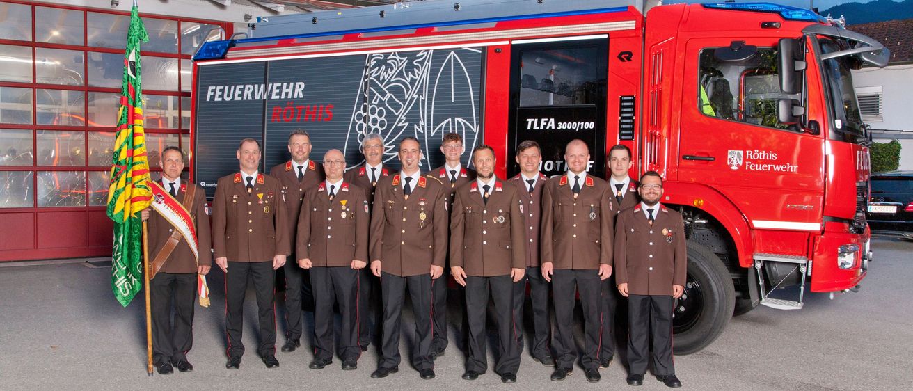 Eine Gruppe von Feuerwehrleuten posiert vor einem roten Feuerwehrauto. Sie sind alle in Uniform gekleidet. Das Wort 'Feuerwehr' ist auf dem Auto geschrieben.