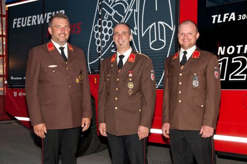 Drei Männer in Militäruniformen stehen vor einem roten Fahrzeug mit einem Design an der Seite. Sie haben alle Medaillen auf der Brust und lächeln.