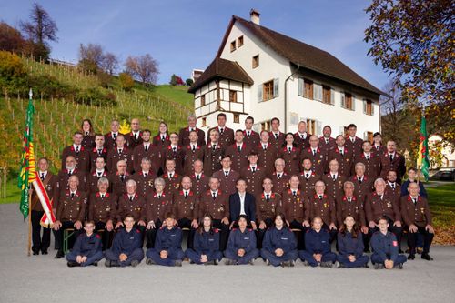 Eine Gruppe Feuerwehrleute in Uniform posiert für ein Foto vor einem Haus mit braunem Dach. Sie stehen und sitzen auf einer Betonfläche.