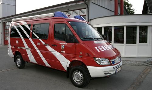 Ein roter Feuerwehrwagen mit weißen Streifen ist vor einem Gebäude geparkt. Der Schriftzug 'FEUERWEHR' steht auf der Seite.