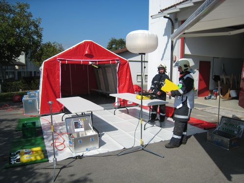 Zwei Feuerwehrleute stehen vor einem roten Zelt und einem Tisch mit Ausrüstung. Einer hält eine gelbe Mappe, während der andere die Ausrüstung auf dem Tisch betrachtet.