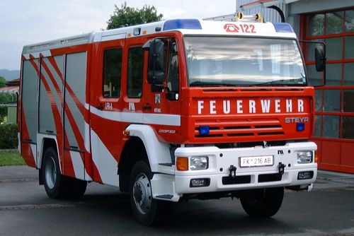 Ein rot-weißer Feuerwehrwagen ist draußen geparkt. Die Front des Fahrzeugs ist mit '122' nummeriert und trägt die Aufschrift 'FEUERWEH' mit dem Nummernschild 'FK 216 AB'.