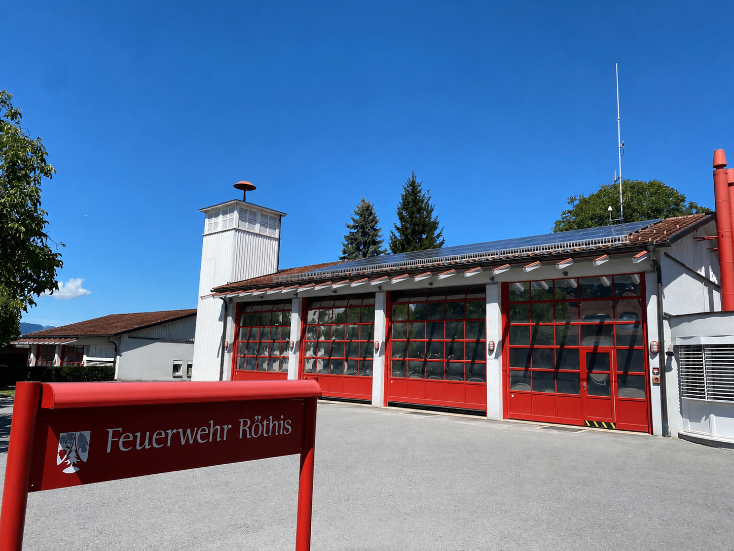 Ein rotes Feuerwehrhaus mit Glastüren und Solarpanelen auf dem Dach. Das Gebäude hat einen Turm und ist von Bäumen umgeben.