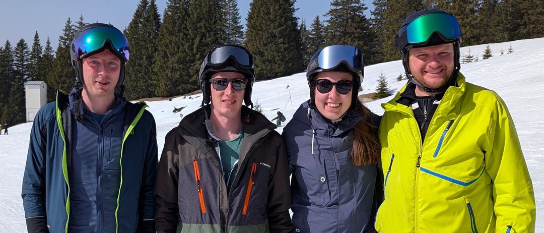 Fünf Personen in Winterkleidung stehen auf einem verschneiten Hang. Drei tragen Skier, einer hält Skistöcke und einer trägt ein rotes Jackett. Im Hintergrund stehen Nadelbäume.