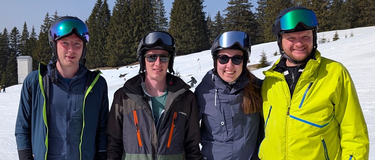 Fünf Personen in Winterkleidung stehen auf einem verschneiten Hang. Drei tragen Skier, einer hält Skistöcke und einer trägt ein rotes Jackett. Im Hintergrund stehen Nadelbäume.