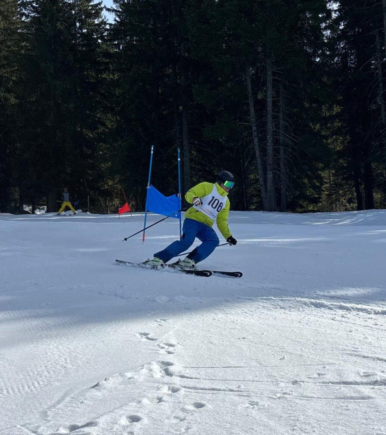 Ein Skifahrer in einem gelben und grünen Outfit mit der Nummer 106 fährt auf einer verschneiten Piste. Es gibt blaue Stangen im Schnee.
