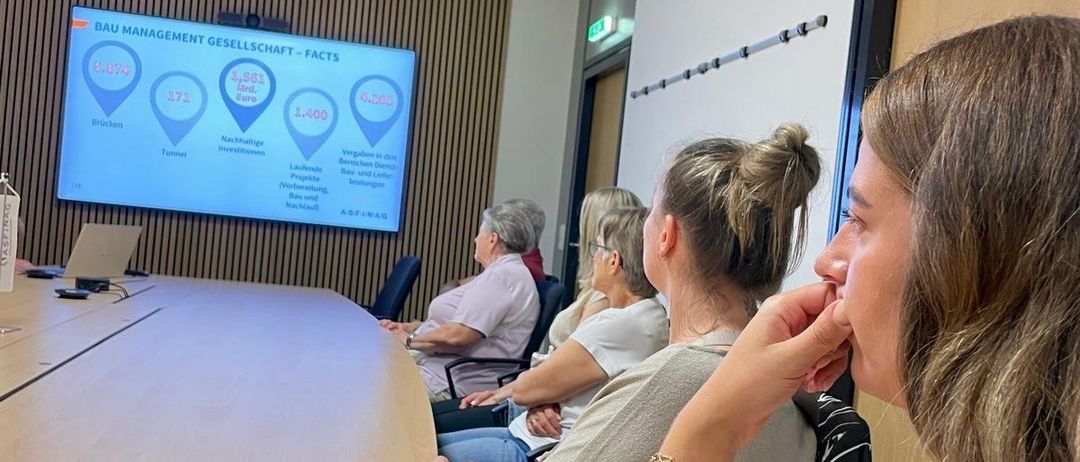 Eine Gruppe von Menschen sitzt in einem Konferenzraum mit einem großen Bildschirm, der Daten anzeigt. Eine Person im Vordergrund hört aufmerksam zu, während andere hinter ihnen sitzen.