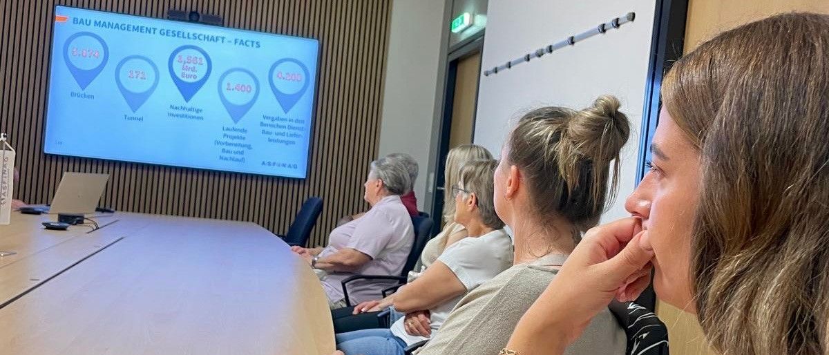 Eine Gruppe von Menschen sitzt in einem Konferenzraum mit einem großen Bildschirm, der Daten anzeigt. Eine Person im Vordergrund hört aufmerksam zu, während andere hinter ihnen sitzen.