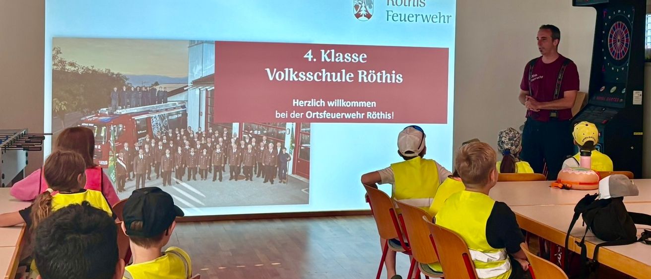 Schüler in gelben Westen sitzen auf Stühlen vor einer Projektionsleinwand, die eine Willkommensnachricht für die 4. Klasse zeigt. Ein Mann in einem burgunderfarbenen Hemd steht hinter der Leinwand.
