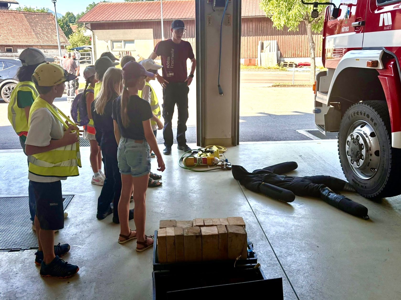 Eine Gruppe von Kindern beobachtet eine Feuerwehrübung in einer Garage. Eine Puppe liegt auf dem Boden, während der Feuerwehrmann den Kindern etwas erklärt.