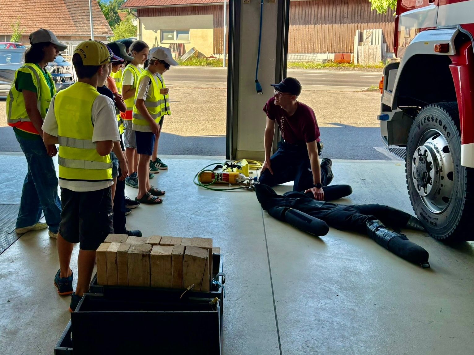 Ein Feuerwehrmann demonstriert in einer Garage vor einer Gruppe von Kindern mit Warnwesten, wie man einen Schutzanzug anlegt.