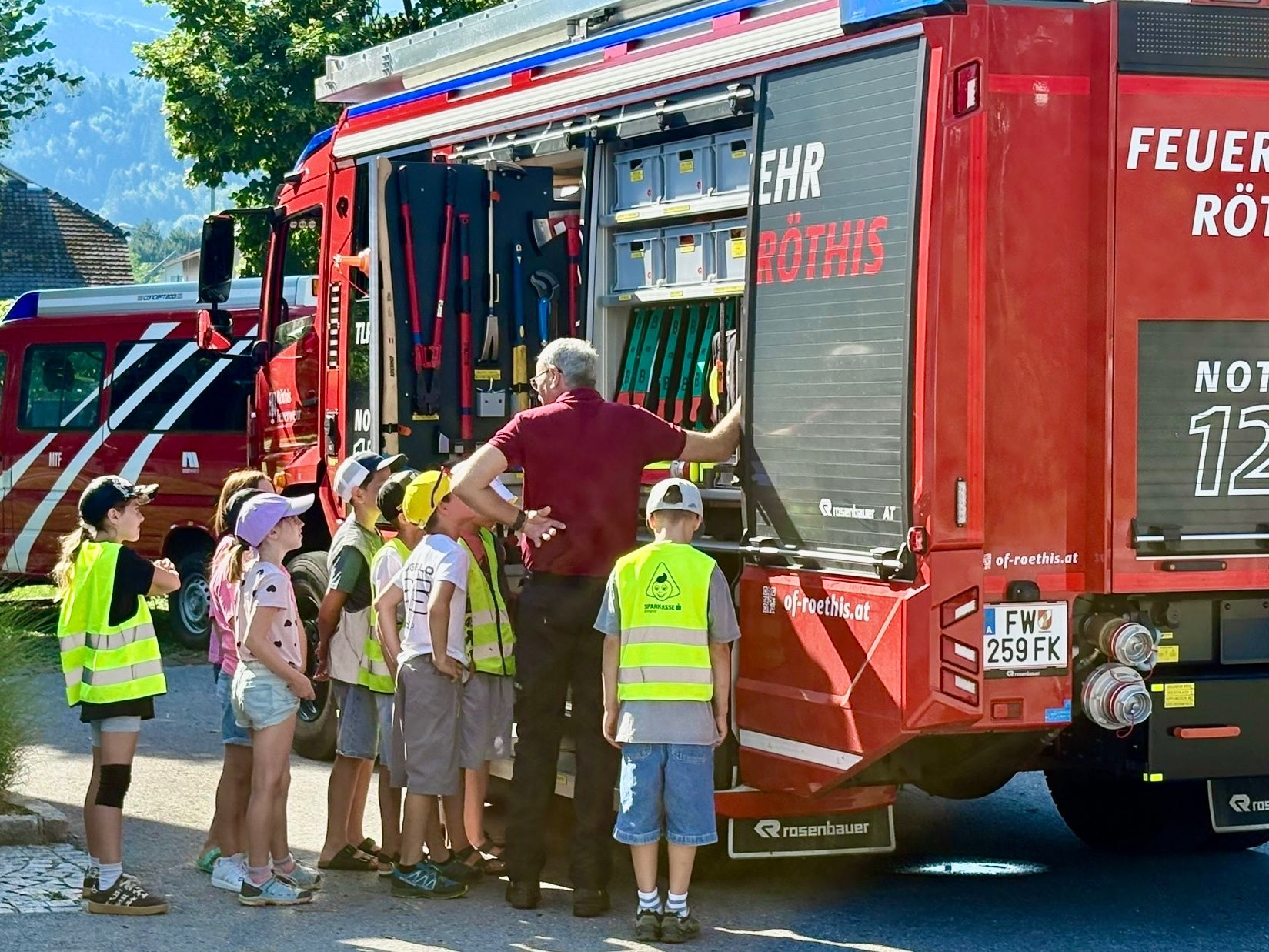 Eine Gruppe von Kindern und Erwachsenen steht um einen roten Feuerwehrwagen mit dem Wort Ehr Rothis an seiner Seite. Ein Mann zeigt auf die Ausrüstung im Inneren.