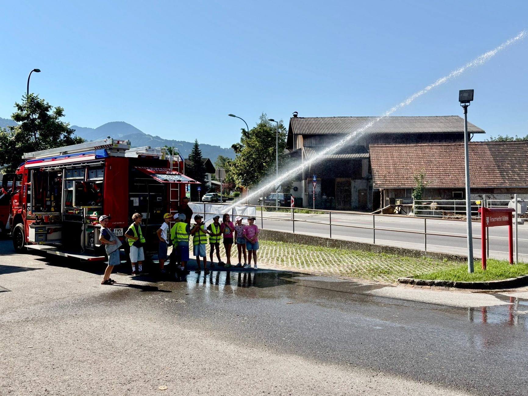 Ein Feuerwehrauto sprüht Wasser, während eine Gruppe von Menschen in Westen und Hüten in der Nähe steht. Hinter ihnen sind Gebäude und ein Metallzaun. Bäume und Berge sind im Hintergrund sichtbar.