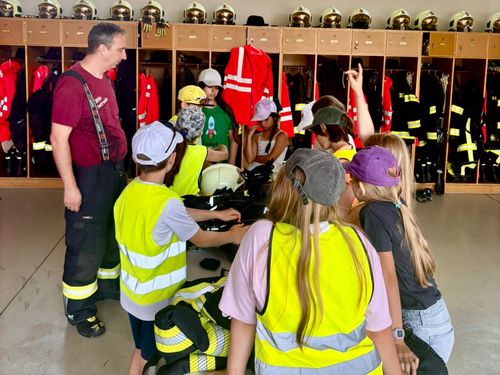 Ein erwachsener Mann spricht mit Kindern in Sicherheitswesten in einer Feuerwache. Einige Kinder sitzen, andere stehen, während Helme und Jacken in Spinden hängen.