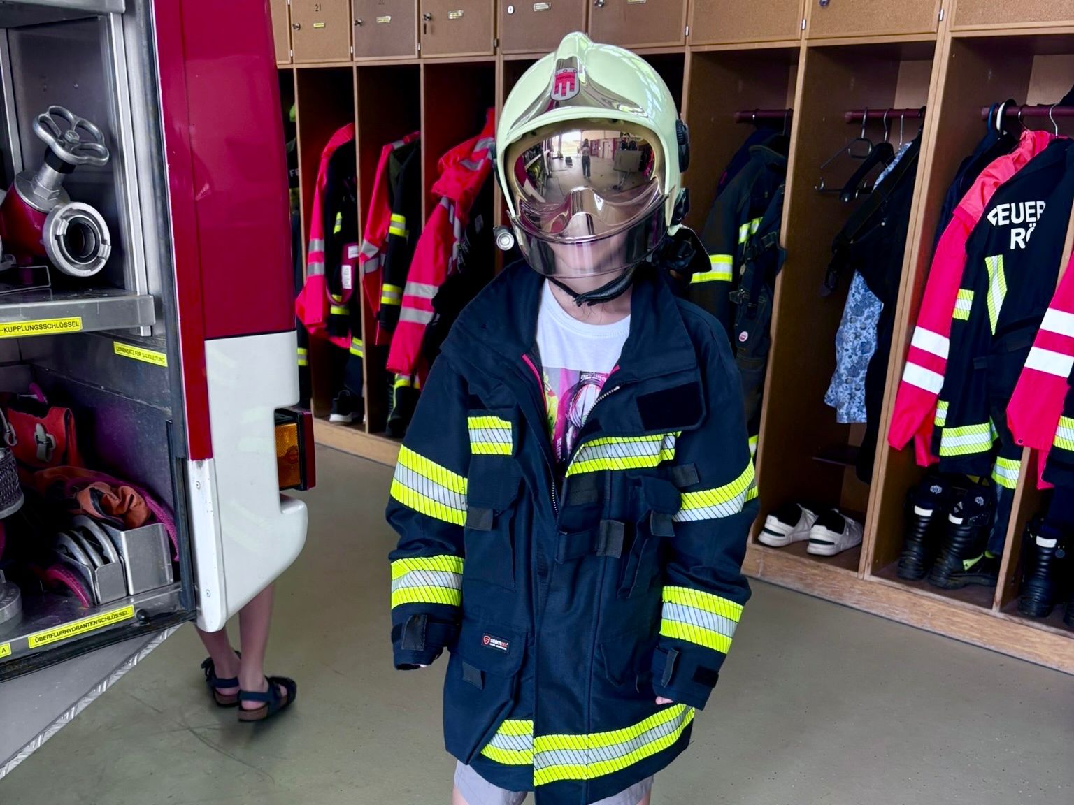 Eine junge Person in einer Feuerwehruniform steht in einem Umkleideraum neben einem Feuerwehrauto. Die Uniform besteht aus einem reflektierenden Helm und einer Jacke mit gelben und roten Streifen.