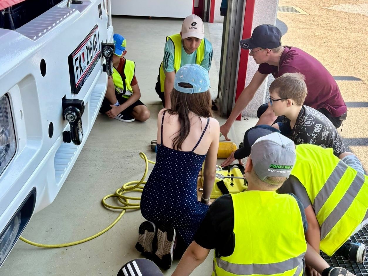 Eine Gruppe von Menschen in reflektierenden Westen ist in einer Garage um ein Fahrzeug versammelt. Sie scheinen mit einer Aufgabe beschäftigt zu sein, wobei sich eine Person beugt und andere stehen.