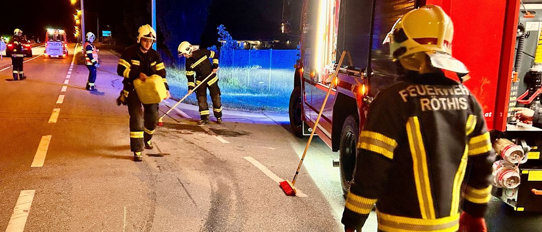 Feuerwehrleute reinigen die Straße bei Nacht, ein Feuerwehrauto steht in der Nähe. Einer hält eine Kehrschaufel, ein anderer trägt einen gelben Beutel.