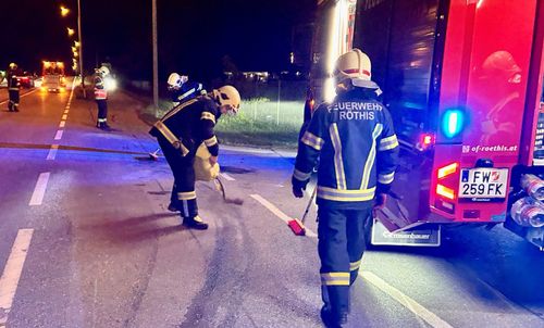 In der Nacht putzen zwei Feuerwehrleute die Straße, einer mit einem Besen und der andere mit einer Schaufel. Der Text 'Feuerwehr Rothis' steht auf ihren Uniformen.