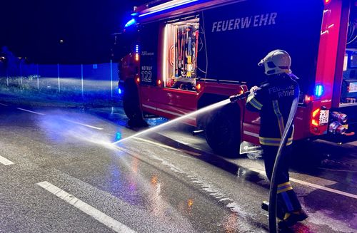 Ein Feuerwehrmann sprüht Wasser aus einem Feuerwehrauto auf einer nassen Straße bei Nacht. Das Fahrzeug ist mit 'FEUERWEHR' und 'NOTRUF 122' gekennzeichnet.