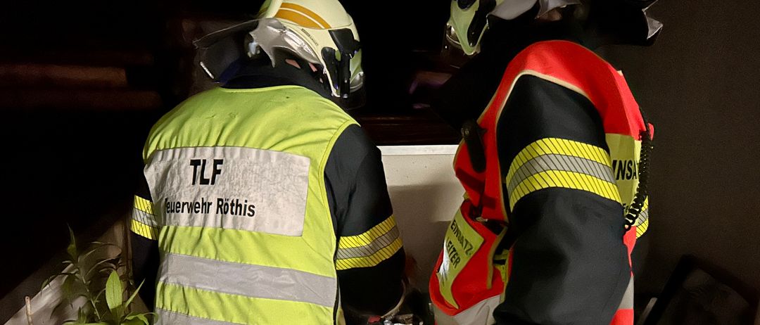 Zwei Feuerwehrleute in reflektierenden Sicherheitsjacken und Helmen stehen drinnen, einer hält eine brennende Laterne. Auf der linken Jacke stehen 'TLF' und 'Feuerwehr Rothis'.