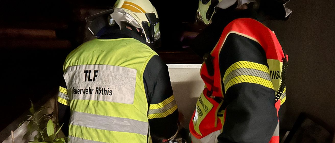 Zwei Feuerwehrleute in reflektierenden Sicherheitsjacken und Helmen stehen drinnen, einer hält eine brennende Laterne. Auf der linken Jacke stehen 'TLF' und 'Feuerwehr Rothis'.