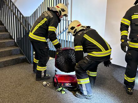 Zwei Feuerwehrleute, die Helme und Uniformen mit gelben und schwarzen Streifen tragen, arbeiten an einem roten tragbaren Ventilator auf dem Teppichboden.