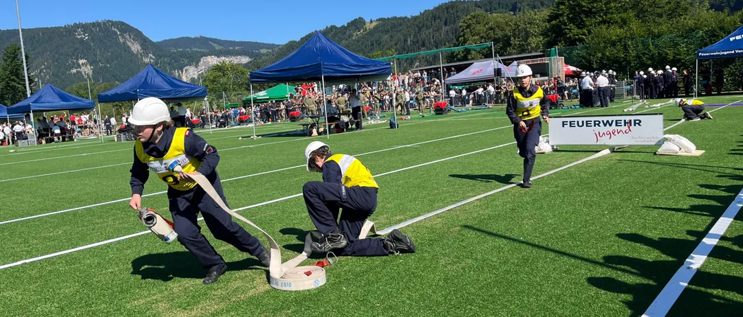 Drei Feuerwehrleute üben auf einem Feld, während eine Menge zuschaut. Einer zieht einen Schlauch, während andere sich beugen, um ihn zu verlegen. Berge und Bäume sind im Hintergrund.