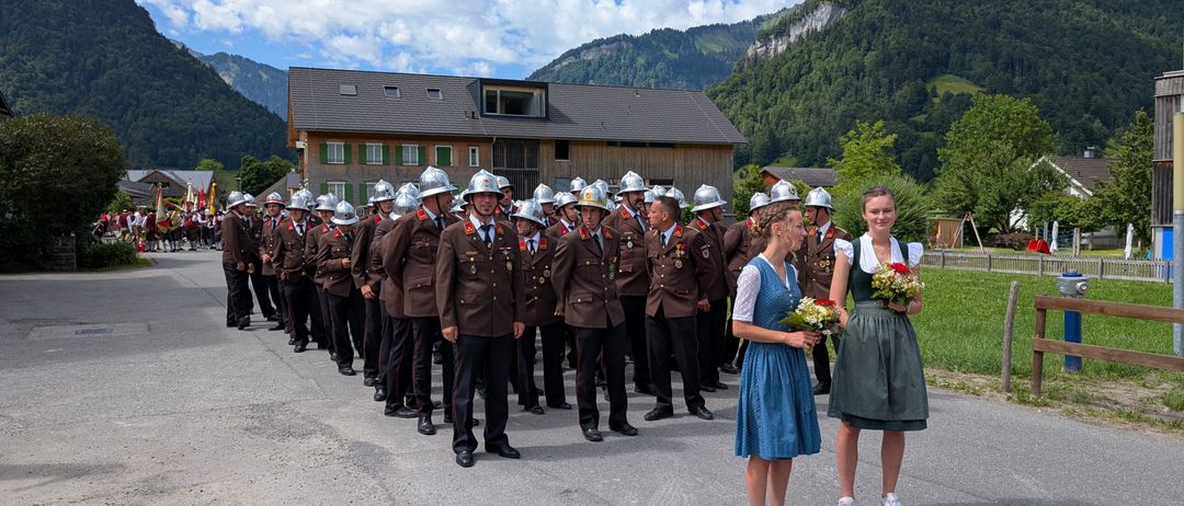 Eine Gruppe uniformierter Personen, möglicherweise Feuerwehrleute, stehen in einer Reihe auf einer Straße mit Bergen im Hintergrund. Zwei Frauen mit Blumen stehen in der Nähe.