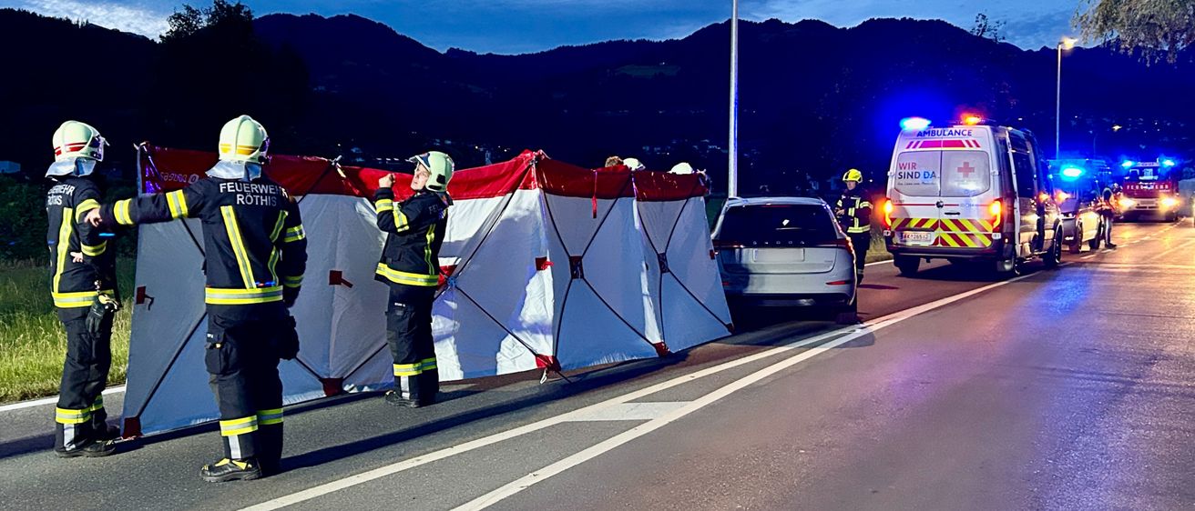 Rettungskräfte richten auf einer Straße ein Zelt auf, mit Bergen im Hintergrund. Ein Krankenwagen mit Warnleuchten ist am Straßenrand geparkt.