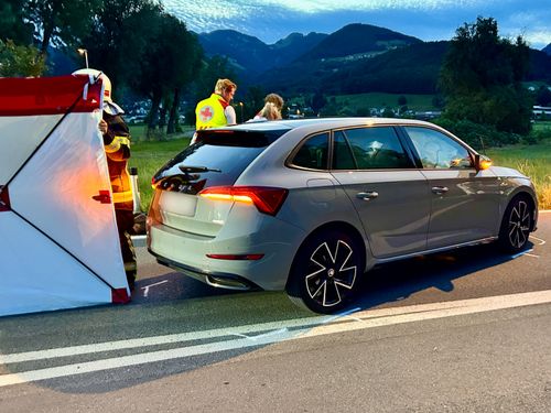 Eine Person in einer reflektierenden Weste steht neben einem Auto am Straßenrand, möglicherweise ein Feuerwehrmann. Das Auto ist auf der Straße geparkt.