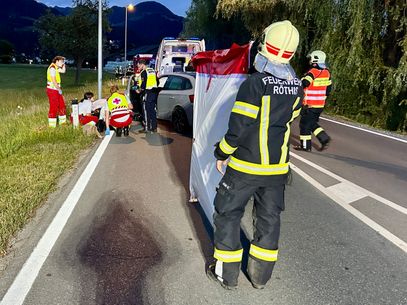 Rettungskräfte kümmern sich um verletzte Personen am Straßenrand. Ein Feuerwehrmann steht mit einem Schutzzelt bereit. Ein Autounfall ist wahrscheinlich.