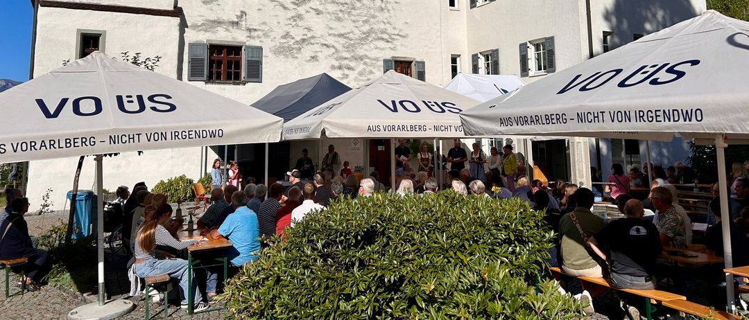 Eine Gruppe von Menschen versammelt sich unter Schirmen vor einem großen weißen Gebäude mit brauner Einfassung. Das Gebäude hat viele Fenster und ein braunes Dach. Die Menschen sitzen an Tischen und unterhalten sich.