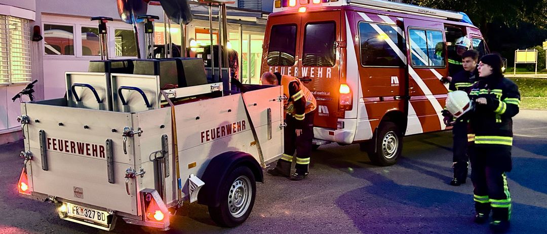 Ein Feuerwehrauto mit blinkenden Lichtern ist vor einem Gebäude geparkt. Ein Feuerwehrmann arbeitet an der Ausrüstung auf dem Anhänger.