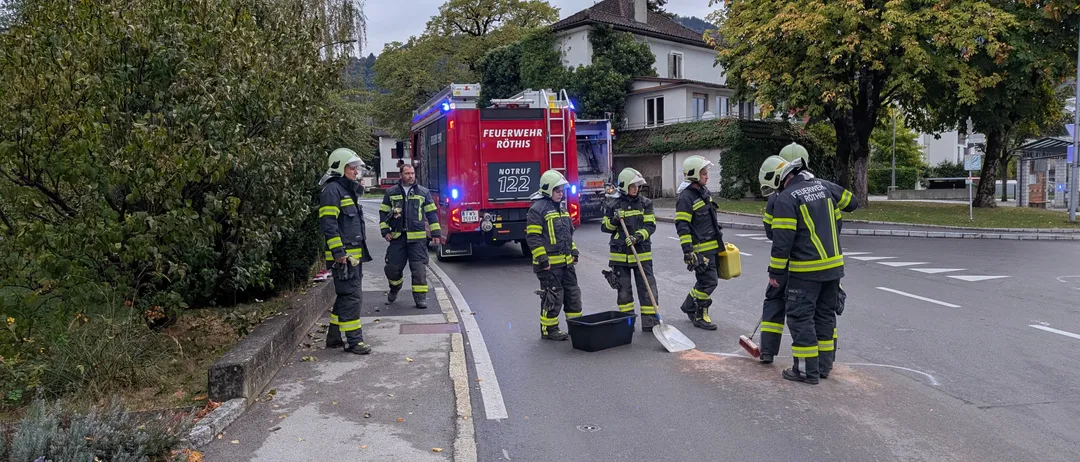 Eine Gruppe von Feuerwehrleuten steht am Straßenrand, einer von ihnen hält eine Schaufel und einen Eimer. Sie tragen vollständige Schutzausrüstung, einschließlich Helme und Stiefel. Ein Feuerwehrwagen ist in der Nähe geparkt, und es gibt Bäume und ein Haus im Hintergrund.