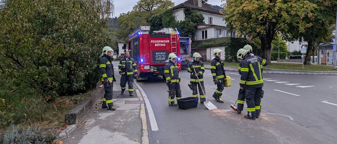 Eine Gruppe von Feuerwehrleuten steht am Straßenrand, einer von ihnen hält eine Schaufel und einen Eimer. Sie tragen vollständige Schutzausrüstung, einschließlich Helme und Stiefel. Ein Feuerwehrwagen ist in der Nähe geparkt, und es gibt Bäume und ein Haus im Hintergrund.