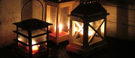Drei Laternen sind beleuchtet und gegen eine Wand gestellt, die ein warmes Licht verbreiten. Jede Laterne hat ein eigenes Design und Größe, wobei die größte ein Kreuzmuster aufweist.